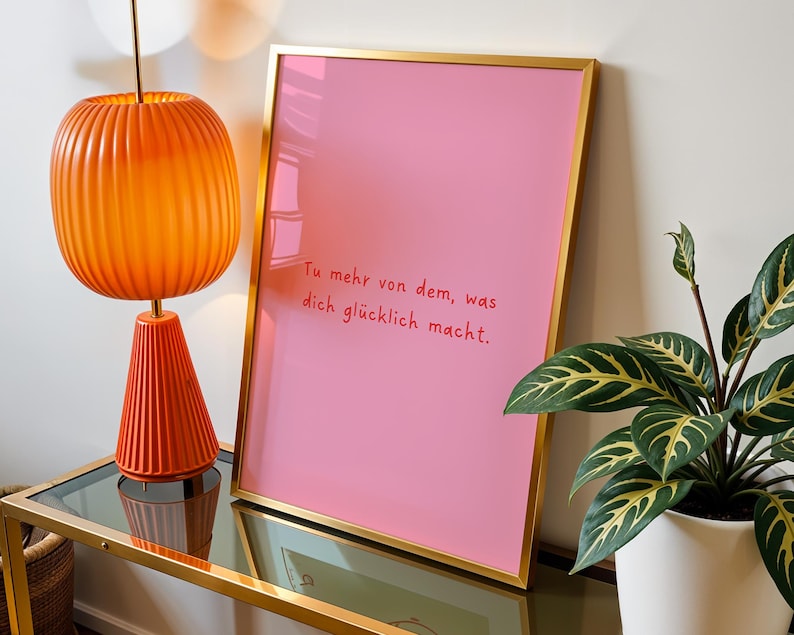May include: A framed pink print with gold trim displays the text "Tu mehr von dem, was dich gl&uuml;cklich macht." An orange lamp with a ribbed shade and base sits on a glass-topped table. A potted plant with green and yellow leaves is also visible.