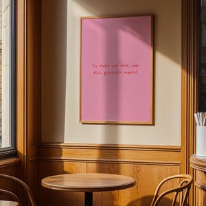 May include: A framed pink poster with the text "Tu mehr von dem, was dich gl&uuml;cklich macht." The poster is in a wooden frame and hangs on a beige wall. The image is taken in a cafe setting with wooden tables and chairs.