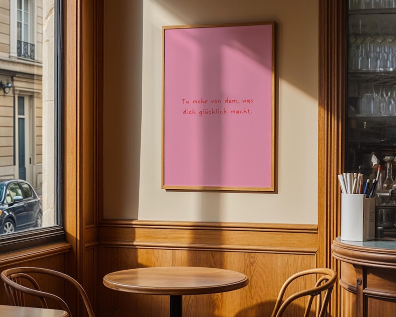 May include: A framed pink poster with the text "Tu mehr von dem, was dich gl&uuml;cklich macht." The poster is in a wooden frame and hangs on a beige wall. The image is taken in a cafe setting with wooden tables and chairs.