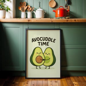 Könnte beinhalten: Ein gerahmter Druck mit dem Text "AVOCUDDLE TIME" über einer Cartoon-Illustration von zwei sich umarmenden Avocados. Die Avocados sind grün mit braunen Kernen und lächelnden Gesichtern. Der Rahmen ist schwarz.