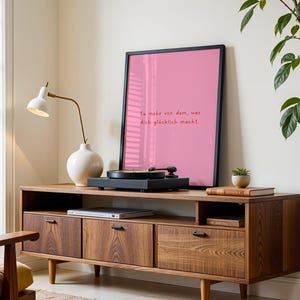 May include: A mid-century modern style wooden media console with a record player, framed pink print, and a white lamp. The pink print has the text "Tu mehr von dem, was dich gl&uuml;cklich macht."
