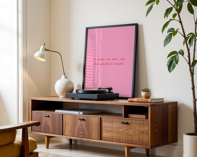 May include: A mid-century modern style wooden media console with a record player, framed pink print, and a white lamp. The pink print has the text "Tu mehr von dem, was dich gl&uuml;cklich macht."