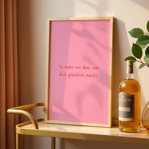 May include: A framed pink print with the German text "Tu mehr von dem, was dich gl&uuml;cklich macht." A bottle of liquor and a vase with greenery sit on a gold table. A beige curtain is visible on the left.