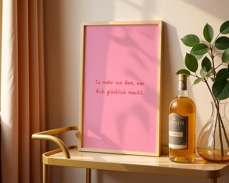 May include: A framed pink print with the German text "Tu mehr von dem, was dich gl&uuml;cklich macht." A bottle of liquor and a vase with greenery sit on a gold table. A beige curtain is visible on the left.