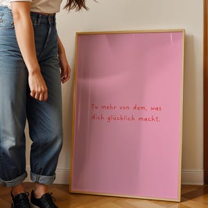May include: A framed pink poster with the German text "Tu mehr von dem, was dich gl&uuml;cklich macht." in red script. The poster has a light wood frame and is leaning against a wall.