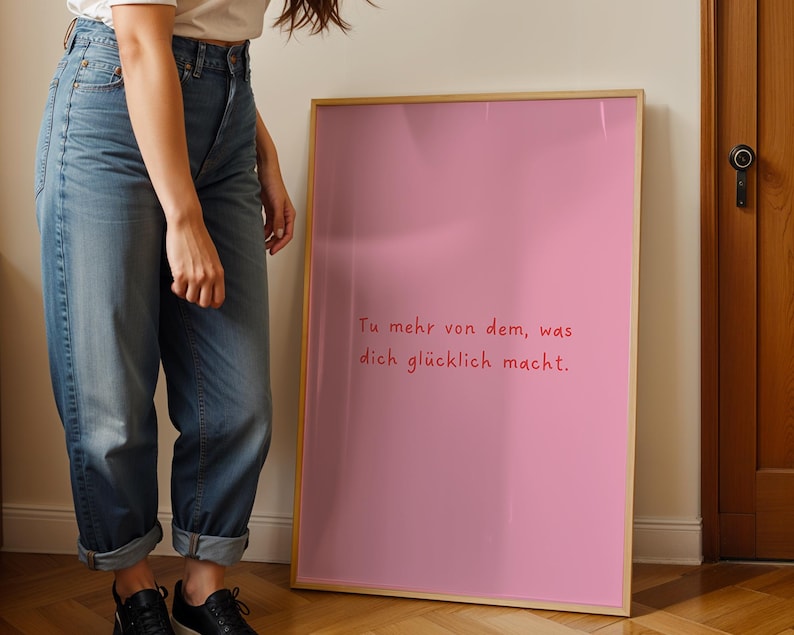 May include: A framed pink poster with the German text "Tu mehr von dem, was dich gl&uuml;cklich macht." in red script. The poster has a light wood frame and is leaning against a wall.