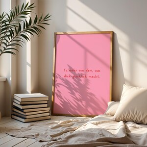 May include: A framed pink print with the text "Tu mehr von dem, was dich gl&uuml;cklich macht." A stack of books and a potted plant are in the foreground. A beige pillow and blanket are in the lower right corner. The print is leaning against a wall.