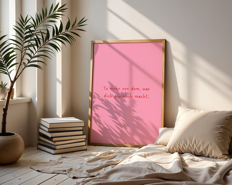 May include: A framed pink print with the text "Tu mehr von dem, was dich gl&uuml;cklich macht." A stack of books and a potted plant are in the foreground. A beige pillow and blanket are in the lower right corner. The print is leaning against a wall.