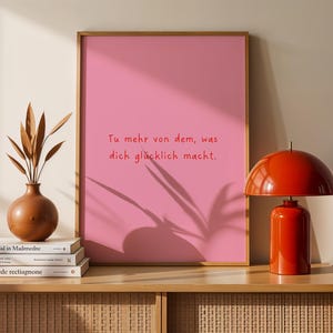 May include: A framed pink print with the text "Tu mehr von dem, was dich gl&uuml;cklich macht." A red lamp, a brown vase with dried plants, and stacked books are on a wooden cabinet.
