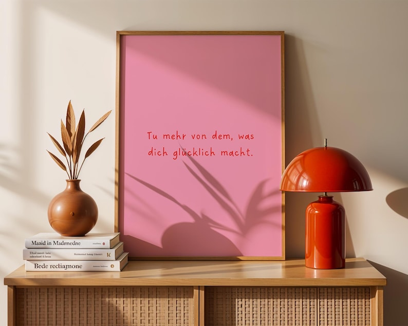 May include: A framed pink print with the text "Tu mehr von dem, was dich gl&uuml;cklich macht." A red lamp, a brown vase with dried plants, and stacked books are on a wooden cabinet.