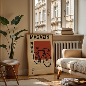 May include: A framed poster with the word "MAGAZIN" and Japanese characters, featuring a black bicycle illustration on a red and beige background. The poster is displayed in a living room with a beige sofa, wooden chair, and a large potted plant.