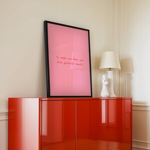May include: A framed pink art print with the text "Tu mehr von dem, was dich gl&uuml;cklich macht" in red lettering. The print is displayed on a glossy red cabinet, with a white lamp and decorative figurine.