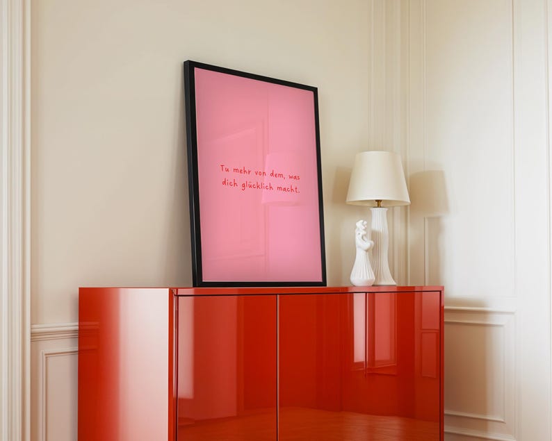 May include: A framed pink art print with the text "Tu mehr von dem, was dich gl&uuml;cklich macht" in red lettering. The print is displayed on a glossy red cabinet, with a white lamp and decorative figurine.