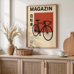May include: A framed poster with a bicycle illustration. The poster has a beige background with a red square and black text that reads "MAGAZIN" and "APRIL 1964 4". A black bicycle is in the center. The poster is displayed on a light wood cabinet.