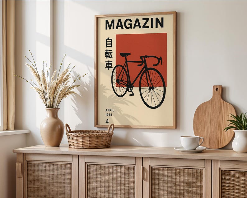 May include: A framed poster with a bicycle illustration. The poster has a beige background with a red square and black text that reads "MAGAZIN" and "APRIL 1964 4". A black bicycle is in the center. The poster is displayed on a light wood cabinet.