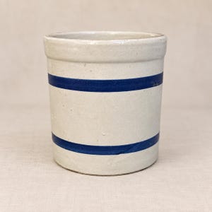 May include: A cylindrical stoneware crock featuring a light beige, off-white finish with two distinct, horizontal cobalt blue stripes encircling the body. The vessel has a thick, reinforced rim and a smooth, matte-textured exterior surface.