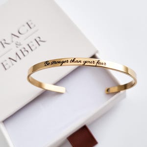 May include: A gold-colored cuff bracelet with the inscription "Be stronger than your fear" in black lettering. The bracelet is displayed inside a white gift box with the words "Grace & Ember" embossed on the lid.