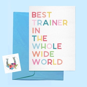 May include: A white greeting card with the text "BEST TRAINER IN THE WHOLE WIDE WORLD" in various colours. It is placed with a blue envelope and a sticker featuring a horseshoe and floral design.