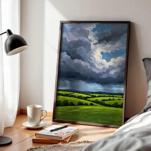 May include: A framed landscape painting depicts a dramatic sky with dark, stormy clouds and a bright patch of blue. The painting shows rolling green hills and a hint of rain. A white coffee cup and a notebook with a pen are in the foreground.