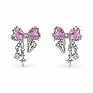 Könnte beinhalten: Ein Paar silberfarbene Ohrringe in Schleifenform mit zwei herzförmigen rosa Edelsteinen in der Mitte. Jeder Ohrring verfügt über hängende Anhänger, bestehend aus einem kleinen klaren Kristall und einem vierzackigen Stern.