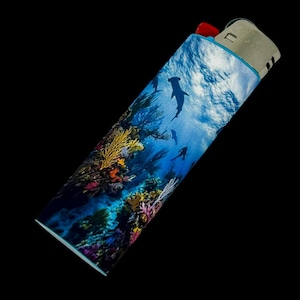 May include: A decorative lighter featuring an underwater scene with hammerhead sharks, coral, and divers. The lighter has a red ignition button and a silver metal top. The design wraps around the lighter, showcasing a vibrant ocean environment.