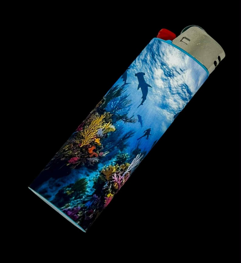 May include: A decorative lighter featuring an underwater scene with hammerhead sharks, coral, and divers. The lighter has a red ignition button and a silver metal top. The design wraps around the lighter, showcasing a vibrant ocean environment.