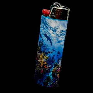 May include: A cylindrical lighter with a vibrant underwater scene featuring dolphins, coral, and a diver. The lighter has a red ignition button and a silver metal top, set against a black background.