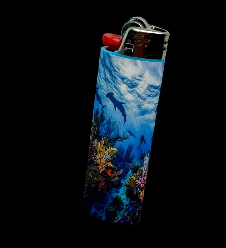 May include: A cylindrical lighter with a vibrant underwater scene featuring dolphins, coral, and a diver. The lighter has a red ignition button and a silver metal top, set against a black background.
