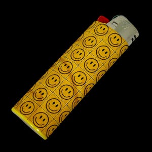May include: A yellow lighter case with a repeating pattern of black smiley faces. The case is rectangular and fits a standard lighter, with a red top peeking out. The background is black.