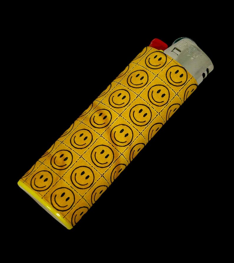 May include: A yellow lighter case with a repeating pattern of black smiley faces. The case is rectangular and fits a standard lighter, with a red top peeking out. The background is black.
