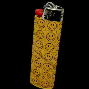 May include: A yellow lighter case with a repeating pattern of black smiley faces. The lighter itself is silver with a red ignition button, set against a black background.