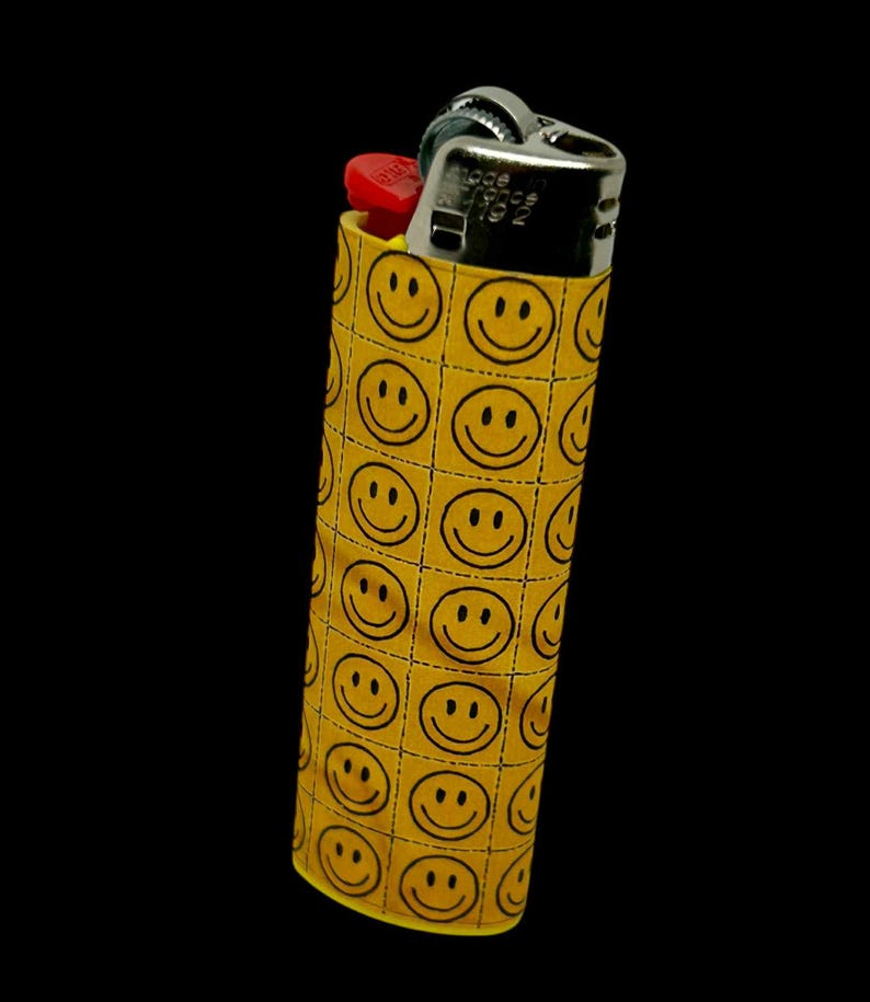 May include: A yellow lighter case with a repeating pattern of black smiley faces. The lighter itself is silver with a red ignition button, set against a black background.