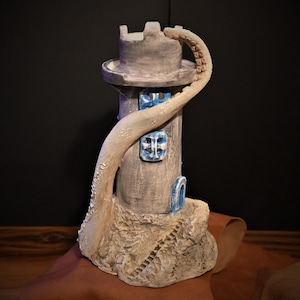 Puede incluir: Una escultura de faro de cerámica gris con detalles de ventanas azules y un tentáculo texturizado blanquecino que lo envuelve. El faro se asienta sobre una base rocosa con una pequeña escalera.