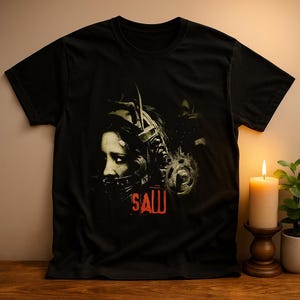 May include: Black t-shirt featuring a distressed graphic from the horror film "Saw." The design includes a close-up of a person wearing a head device, with the word "SAW" in red block letters. A lit candle and plant are in the background.