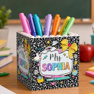 Puede incluir: Un portalápices cuadrado con un patrón de cuaderno de composición en blanco y negro. El frente presenta una etiqueta blanca con el texto Mrs. SOPHIA en letras coloridas, rodeado de ilustraciones de un lazo amarillo, una paleta y un globo.