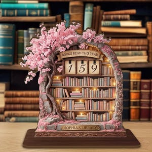 Puede incluir: Figura decorativa de estantería con un cerezo en flor y el texto "BOOKS READ THIS YEAR 159". La estantería en miniatura está llena de libros pequeños y tiene el texto "SOPHIA'S LIBRARY" en la base. La figura está sobre una base de madera.