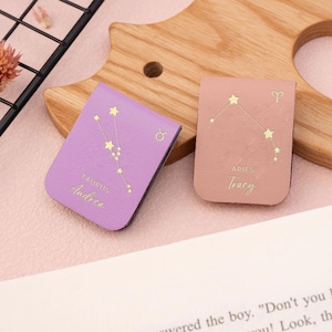 Könnte beinhalten: Zwei Lesezeichen aus Kunstleder, eines in Lavendel und eines in Pfirsich. Das lavendelfarbene Lesezeichen zeigt das Sternbild Stier und den Namen "Andrea". Das pfirsichfarbene Lesezeichen zeigt das Sternbild Widder und den Namen "Tracy". Beide haben goldfarbene Sterndesigns.