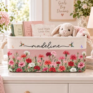 May include: A rectangular fabric storage bin with rope handles, featuring a pink and cream color scheme. The front displays a floral pattern of red, pink, and white carnations, two hummingbirds, and the name "madeline" in black cursive script.