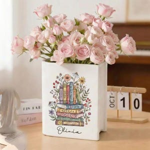 Puede incluir: Un jarrón rectangular blanco lleno de rosas rosas. El jarrón está decorado con una pila de libros coloridos e ilustraciones florales, con el nombre "Olivia" debajo. Un calendario de madera muestra "OCT 10".