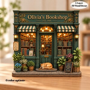 May include: A 5-layer 3D wood decor model of Olivia's Bookshop, featuring a dark green storefront with striped awnings, interior warm lighting, shelves of books, potted plants, a sleeping orange cat, and a sign reading A BOOK IS A JOURNEY THAT NEVER ENDS.
