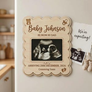 Puede incluir: Un marco de madera con borde festoneado contiene una imagen de ultrasonido. El marco está grabado con "Baby Johnson", "HI MOM HI DAD" y "ARRIVING 20th DECEMBER, 2026 Comming Soon". Una nota que dice "We're expecting!" está adjunta.