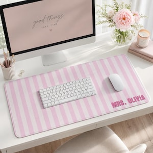 May include: A rectangular desk mat featuring a light pink and white vertical striped pattern. The bottom right corner displays the text MRS. OLIVIA in bold pink lettering. The mat is positioned on a white desk surface beneath a computer keyboard and mouse.
