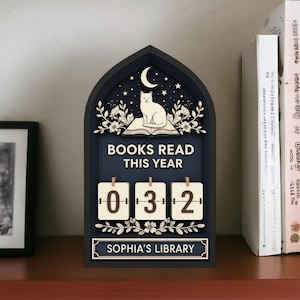 May include: A dark blue wooden bookshelf decor piece with a cat on a book design. The text "BOOKS READ THIS YEAR" is above the number display, which reads "032". The bottom reads "SOPHIA'S LIBRARY".