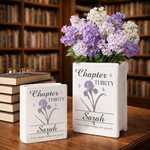 May include: Two white ceramic book-shaped vases with floral designs and the text "Chapter Thirty" and "Sarah." One vase holds a bouquet of purple and white flowers. The vases are on a wooden surface in front of a bookshelf.