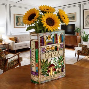 May include: A stained glass vase shaped like a book, filled with sunflowers. The vase is decorated with colorful images of books, flowers, and the name "EMMA". The text "JUST ONE MORE CHAPTER" is also visible.