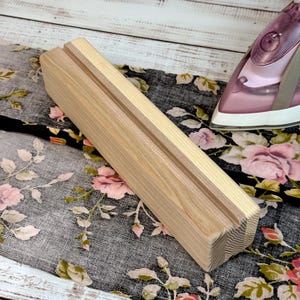 Può includere: Un blocco di legno chiaro con una scanalatura al centro. Il blocco è rettangolare con un bordo smussato. Il blocco è appoggiato su un tessuto a fantasia floreale. Un ferro da stiro rosa è sullo sfondo.
