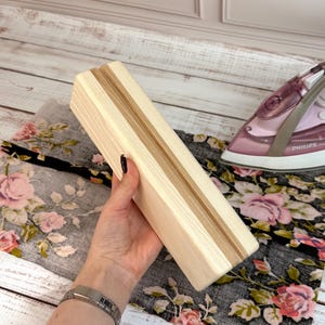 Può includere: Un supporto rettangolare in legno per asse da stiro con una scanalatura al centro. Il supporto è realizzato in legno chiaro con una striscia a contrasto di legno più scuro. Il supporto è tenuto in mano da una persona.