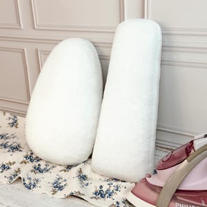 May include: Two white fabric pressing hams and rolls with a smooth texture. One is a rounded, egg-shaped pressing ham, and the other is a long, cylindrical pressing roll. A pink and white steam iron is positioned to the right of the items.