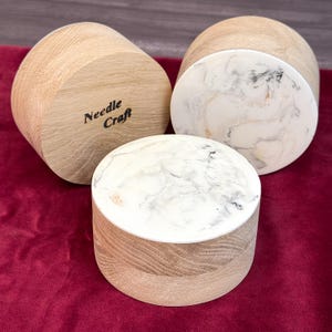 Könnte beinhalten: Drei zylindrische Behälter aus hellem Holz mit glatter Maserung. Zwei Behälter haben weiße Harzoberflächen mit grauen und schwarzen marmorierten Wirbeln. Eine Holzseite ist deutlich mit dem Schriftzug Needle Craft graviert.