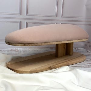 Könnte beinhalten: Ein Bügelbrett aus Holz mit einem hellrosa gepolsterten Oberteil. Das Brett hat eine natürliche Holzoberfläche und ein ovales Design. Der Sockel besteht ebenfalls aus Holz und bietet eine stabile Grundlage für das Bügeln von Kleidung. Ca. 60 cm lang.